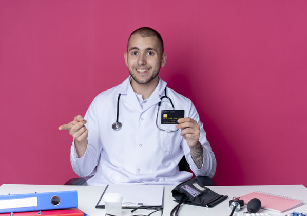桌子面带微笑的年轻男医生穿着医用长袍 听诊器坐在办公桌旁 手里拿着工作工具 笔直地指着粉色的墙上的信用卡微笑粉红色感情