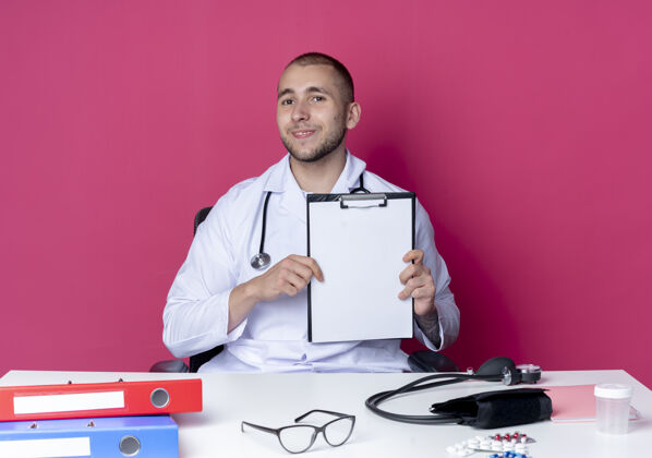 人高兴的年轻男医生穿着医用长袍 听诊器坐在办公桌旁 工作工具上的剪贴板被隔离在粉红色的墙上表演男性长袍