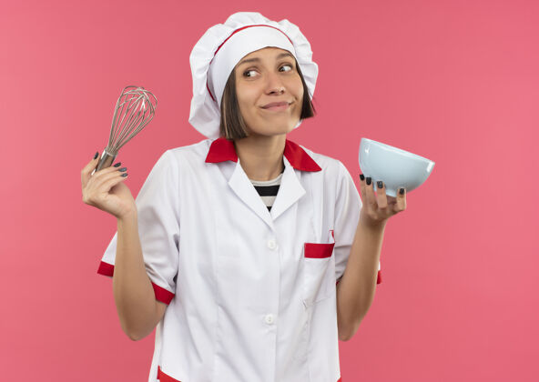 表情年轻的女厨师穿着厨师制服 手里拿着搅拌器和碗 看着粉红色墙上孤立的一面壁板人粉色