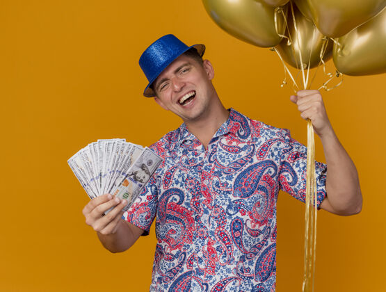 橙色快乐的年轻人戴着蓝色的帽子拿着气球和现金隔离在橙色气球穿现金
