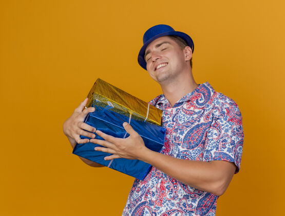 穿上高兴的年轻人闭着眼睛戴着蓝色帽子拥抱礼品盒孤立的橙色眼睛盒子拥抱