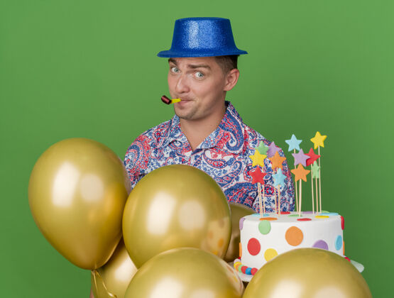 站着高兴的年轻人戴着蓝色的帽子站在气球后面吹蛋糕聚会吹风机隔离在绿色吹年轻人请穿