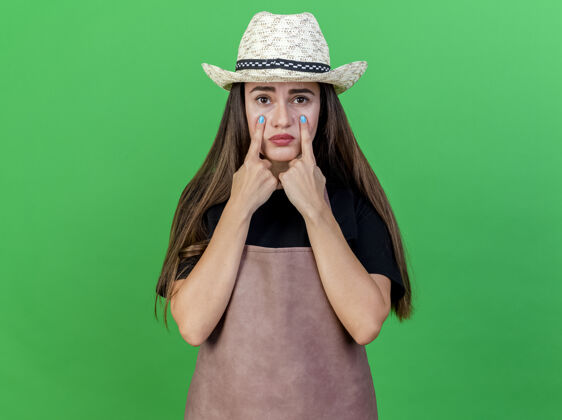 悲伤悲伤美丽的园丁女孩穿着制服戴着园艺帽把手指放在眼睛上孤立的绿色绿色花园手指