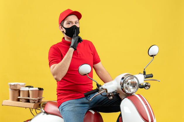 制造俯视图：戴着医用口罩 身穿红色上衣 戴着帽子手套的年轻人坐在踏板车上 在黄色背景上做着沉默的手势车辆红色命令