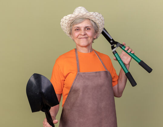 园艺高兴的上了年纪的女园丁戴着园艺帽手持园艺剪刀和铁锹在橄榄绿上帽子黑桃剪刀