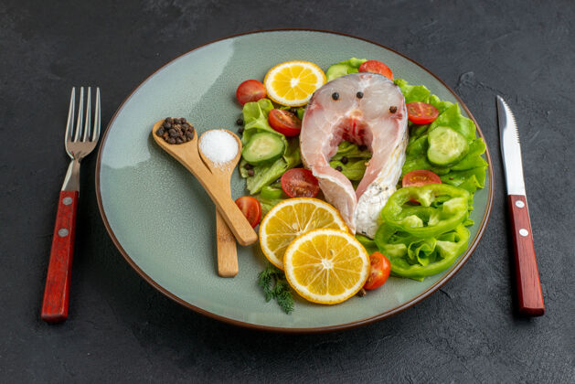 食物侧视图生鱼肉和新鲜切碎的蔬菜柠檬片香料在一个灰色的盘子和餐具设置在黑色苦恼的表面晚餐午餐柠檬