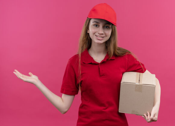 盒子微笑着的年轻送货女孩 穿着红色制服 拿着盒子 在孤立的粉色空间里展示着空手红色制服手
