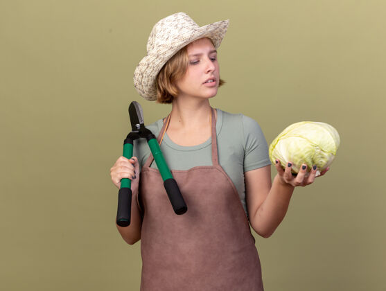 剪刀自信的年轻斯拉夫女园丁戴着园艺帽拿着园艺剪刀看着橄榄绿上的卷心菜园艺卷心菜信心