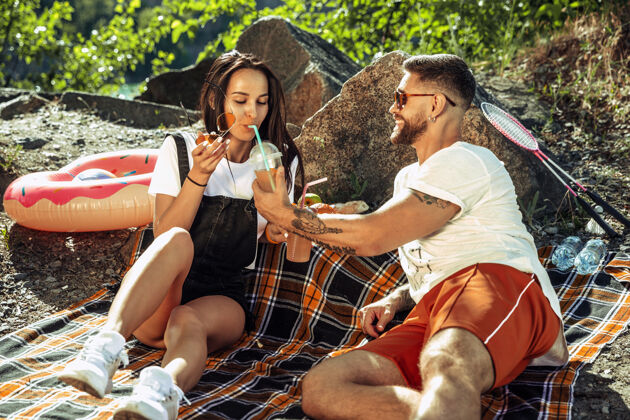 约会一对年轻的情侣在阳光明媚的日子里在河边野餐女人和男人一起在大自然中度过时光玩得开心 吃 玩 笑关系 爱情 夏天 周末的概念蜜月男孩野餐
