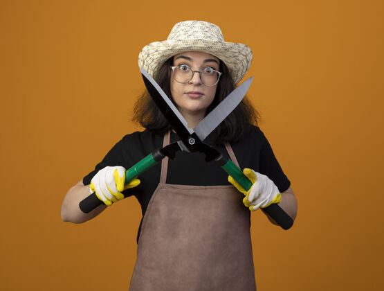 帽子年轻的深色皮肤的女园丁戴着眼镜 穿着制服 戴着园艺帽和手套 手里拿着园艺剪子 隔离在橙色的墙上 留着复印空间年轻花园制服