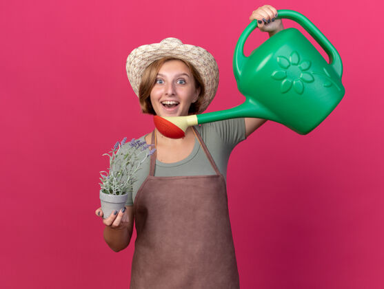 年轻兴奋的年轻斯拉夫女园丁戴着园艺帽拿着浇水罐和粉色花盆里的花花花园园艺