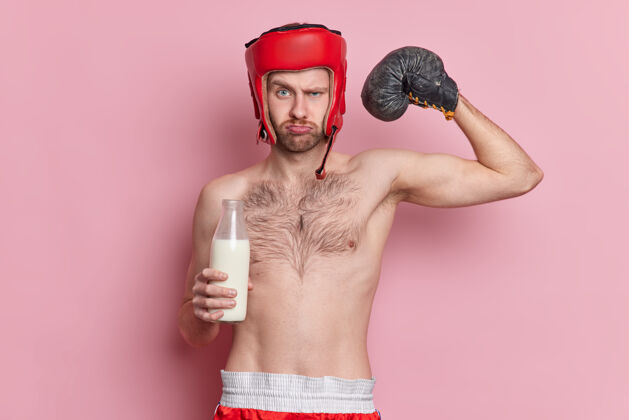 男人严肃的男子拳击手 瘦身 戴拳击手套 戴拳击帽 举起手臂 展示肌肉 喝牛奶 拥有强壮的肱二头肌 展示他的力量和力量运动和激励理念室内粉色比赛