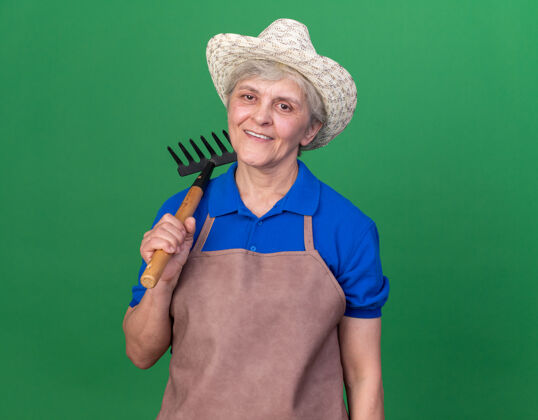 肩膀微笑的上了年纪的女园丁戴着园艺帽 肩上扛着耙子站在草地上老人花园园艺