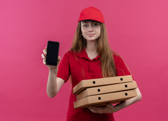 手持穿着红色制服 拿着包裹 在孤立的粉色空间里展示手机的年轻送货女孩很高兴盒子红色请