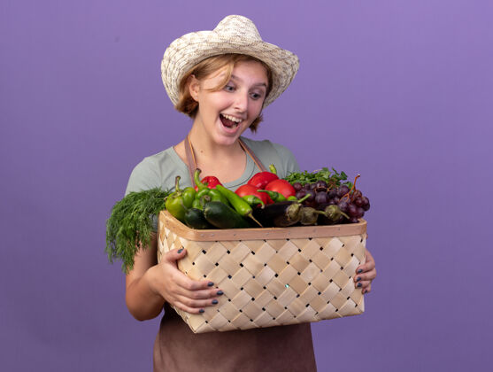 篮子年轻的斯拉夫女园丁惊讶地戴着园艺帽 拿着紫色的菜篮看着斯拉夫花园蔬菜
