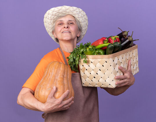 篮子高兴的上了年纪的女园丁戴着园艺帽 手里拿着南瓜和紫菜篮花园南瓜帽子