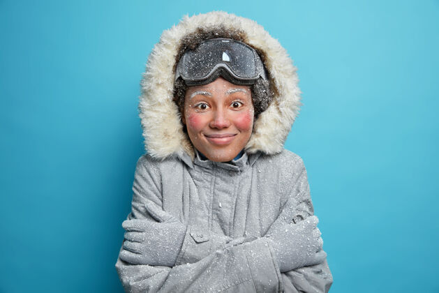 休息寒假和休闲概念开朗的冻僵女人在山上滑雪后感到寒冷颤抖和拥抱自己温暖穿灰色夹克带风帽手套感到满意护目镜女性皮毛