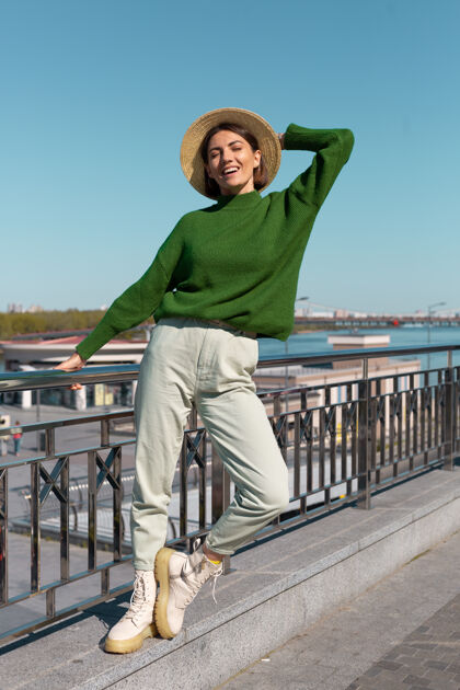 户外穿着绿色休闲毛衣 戴着帽子的时尚女士在户外的桥上欣赏河景 享受夏日阳光明媚的日子阳光梦想欢乐