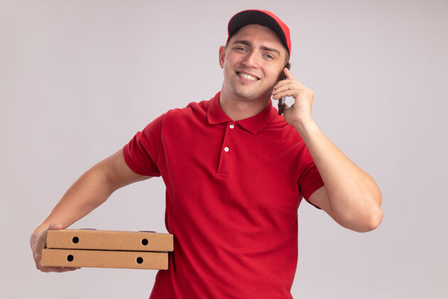 人微笑的年轻送货员身穿制服 戴着帽子 拿着披萨盒 隔着白墙讲电话姿势穿着抱着