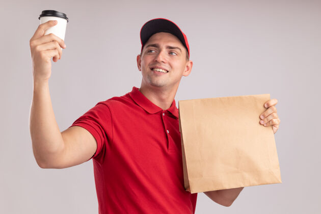 食物面带微笑的年轻送货员穿着制服 戴着帽子 手里拿着纸制食品包 抬头望着隔离在白墙上的一杯咖啡纸递送公民