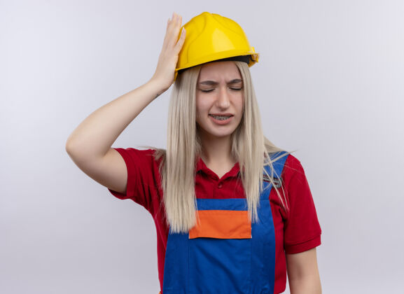 金发年轻的金发碧眼的建筑工女孩 穿着制服 戴着牙套 手放在头上 在孤立的白色空间里闭着眼睛头痛疼痛眼睛年轻