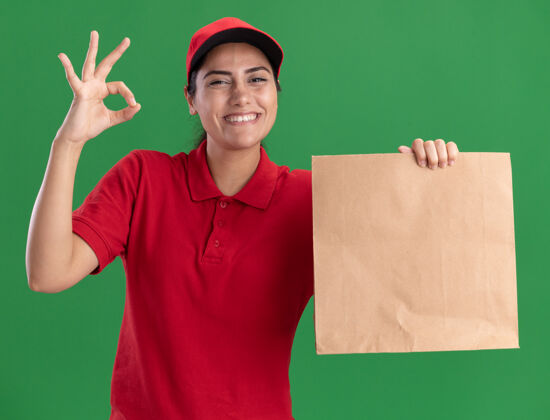 纸带着微笑的年轻送货女孩穿着制服 戴着帽子 拿着纸食品包 在绿色的墙上显示出良好的姿态递送制服姿势