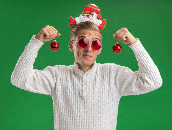 圣诞快乐给人印象深刻的年轻帅哥戴着圣诞老人的头带戴着眼镜拿着圣诞饰品看着隔离在绿色背景上的相机眼镜抱着圣诞老人