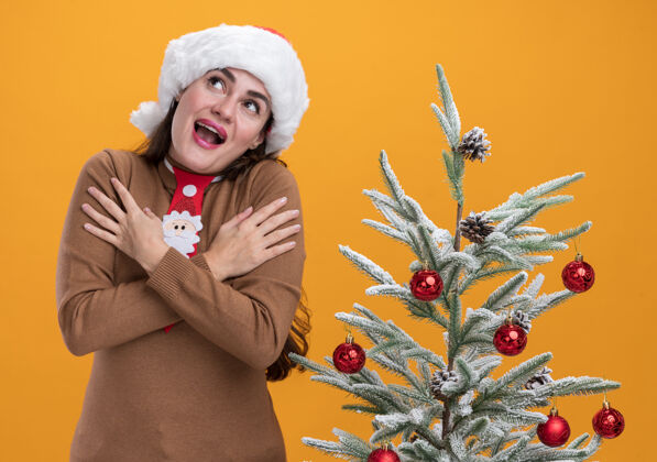 手快乐的仰望着戴着圣诞帽打着领带的年轻漂亮女孩站在圣诞树旁 手放在肩膀上 孤立在橙色的背景上年轻站起来圣诞快乐