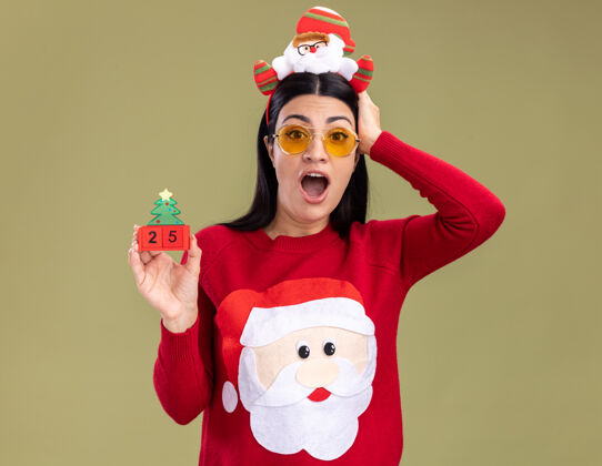 关心关心的年轻白人女孩戴着圣诞老人的头带和带眼镜的毛衣拿着圣诞树玩具和日期保持手在头上看着橄榄绿背景上孤立的相机圣诞老人圣诞快乐日期