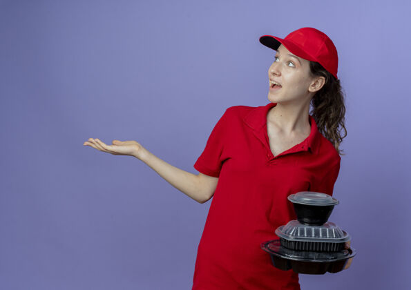 印象令人印象深刻的年轻漂亮的送货女孩穿着红色制服 戴着帽子拿着食品容器看着侧面 显示空的手孤立在紫色背景与复制空间帽子空显示