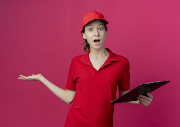 深红色令人印象深刻的年轻漂亮的送货女孩在红色制服和帽子举行剪贴板和显示空的手孤立在深红色的背景举行漂亮剪贴板