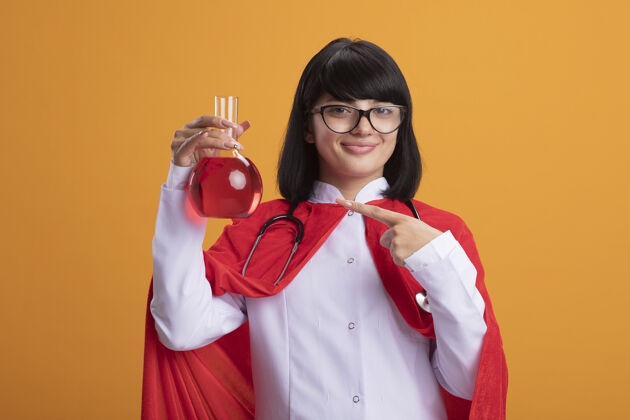 穿年轻的超级英雄女孩戴着听诊器 穿着医用长袍和斗篷 戴着眼镜 指着装满红色液体的化学玻璃瓶点医疗填补