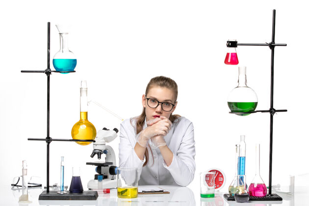 研究前视图穿着白色医疗服的女医生坐在桌子前 用白色背景的解决方案研究病毒大流行化学观白