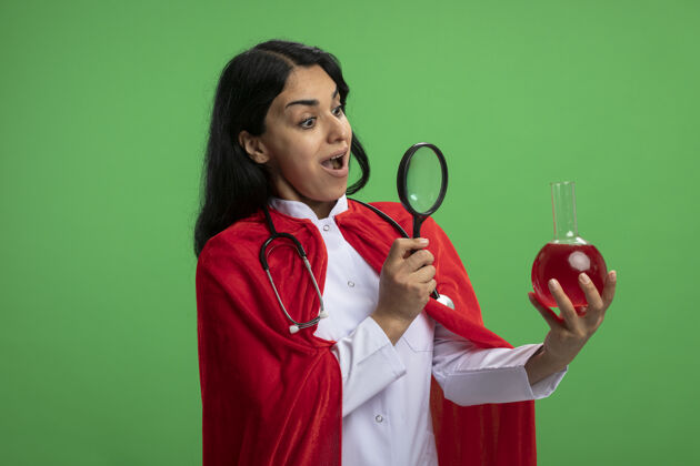 持有惊讶的年轻超级英雄女孩穿着医用长袍 手持听诊器 看着装满红色液体的化学玻璃瓶 放大镜隔离在绿色上化学瓶子惊喜