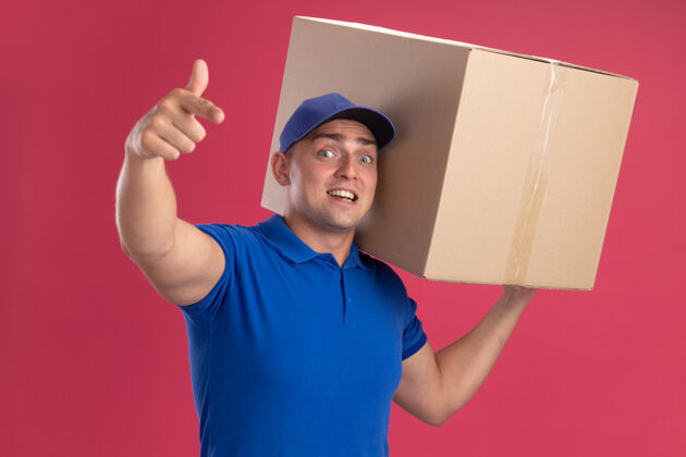 举行惊讶的年轻送货员穿着制服 戴着帽子 肩上扛着一个大箱子 在粉红色的墙上显示着你们的孤立姿态手势穿年轻