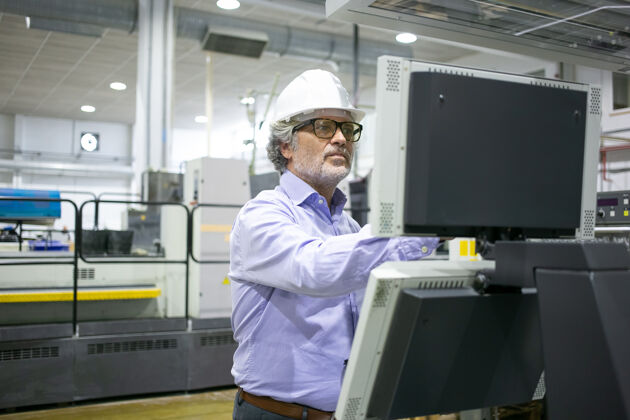 板严肃的男性厂长戴着安全帽和眼镜操作工业机器 按下控制面板上的按钮工作专业工作