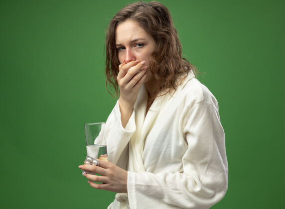 袍咳嗽的年轻病女孩穿着白色长袍拿着一杯水 把手放在嘴上隔离在绿色抱病口