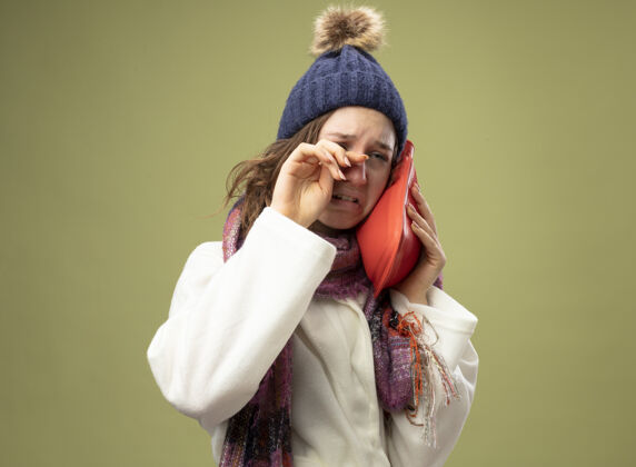 年轻哭泣的生病的年轻女孩穿着白色长袍 戴着冬天的帽子 戴着围巾 把热水袋放在脸颊上 用手在橄榄绿上擦眼睛绿色眼睛湿巾