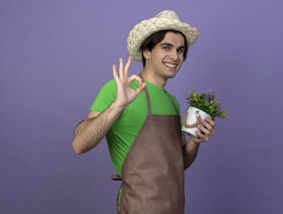 男人微笑的年轻男园丁身着制服 戴着园艺帽 在花盆里捧着鲜花 表现出良好的姿态花盆人穿上