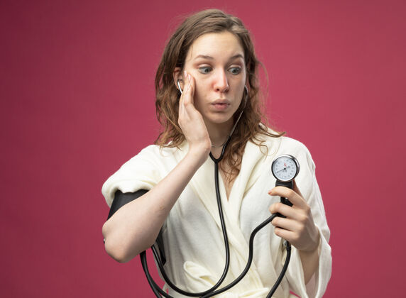 测量惊讶的年轻病女孩穿着白色长袍测量自己的压力血压计把手放在脸颊上隔离粉红色年轻粉色血压计