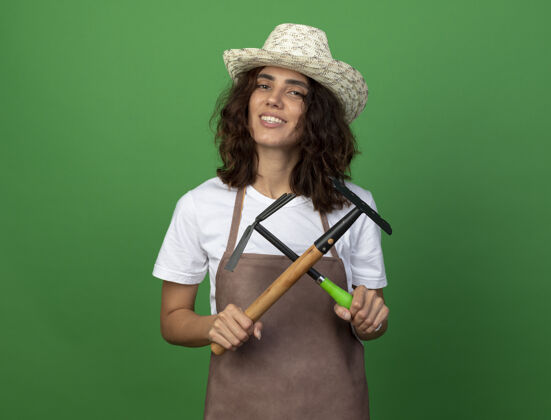 微笑微笑着的年轻女园丁穿着制服戴着园艺帽拿着耙子和锄头耙举行帽子花园