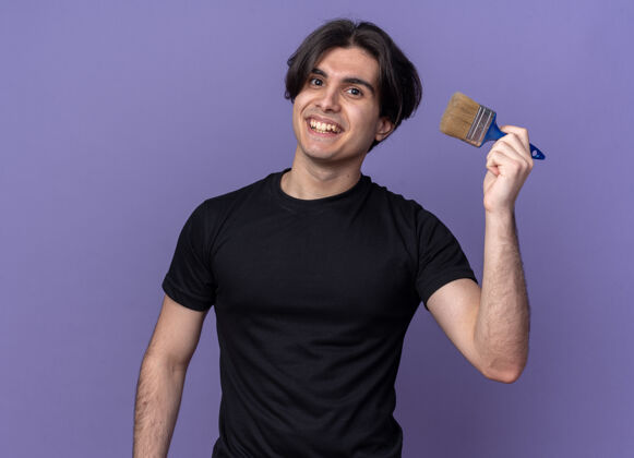 抱着微笑的年轻帅哥穿着黑色t恤拿着油漆刷孤立在紫色的墙上穿微笑小伙子