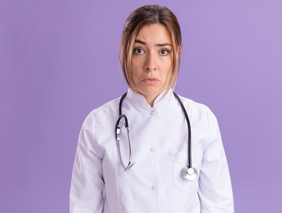 悲伤悲伤的年轻女医生穿着医用长袍 听诊器被隔离在紫色的墙上长袍女性医生