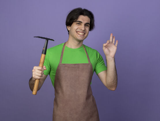 男性微笑的年轻男园丁身着制服 手持锄头耙 表现出良好的姿态好的紫色年轻