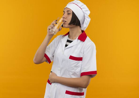 手年轻的女厨师穿着厨师制服 闭上眼睛从杯子里喝水 把手放在肚子上 在橙色的背景上 留有复制空间饮用制服年轻