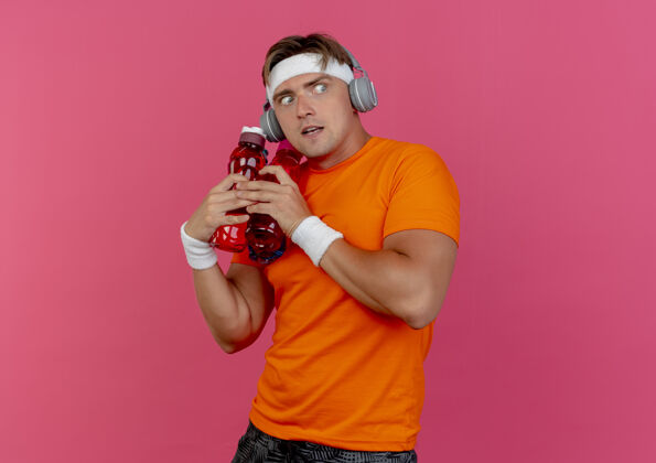 壁板印象深刻的年轻英俊的运动男子戴着头带和腕带和耳机拿着水瓶看着一边孤立的粉红色粉色持有耳机