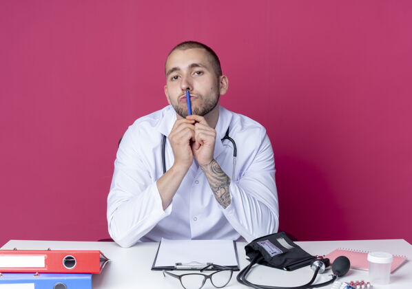 医疗年轻的男医生穿着医用长袍和听诊器坐在办公桌旁 手里拿着笔 用笔触摸嘴唇 看着隔离在粉色背景上的摄像机长袍医生年轻