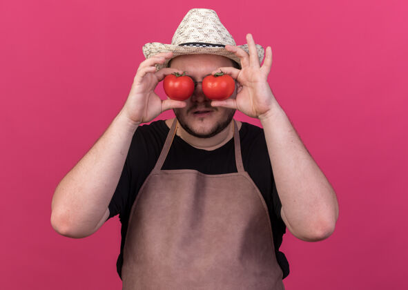 手势高兴的年轻男园丁戴着园艺帽 表现出与隔离在粉红色墙上的西红柿的表情姿态表演请帽子