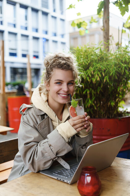 年轻人垂直拍摄了一个美丽的微笑的女人 一边喝着鸡尾酒 一边坐在咖啡馆的户外桌子上 一边用笔记本电脑看着幸福电脑设备笔记本电脑