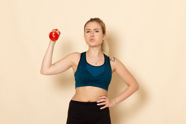 健身正面图身穿运动装的年轻女性手持红色哑铃站在白墙上运动身体健康美容锻炼的女人运动器材女士服装